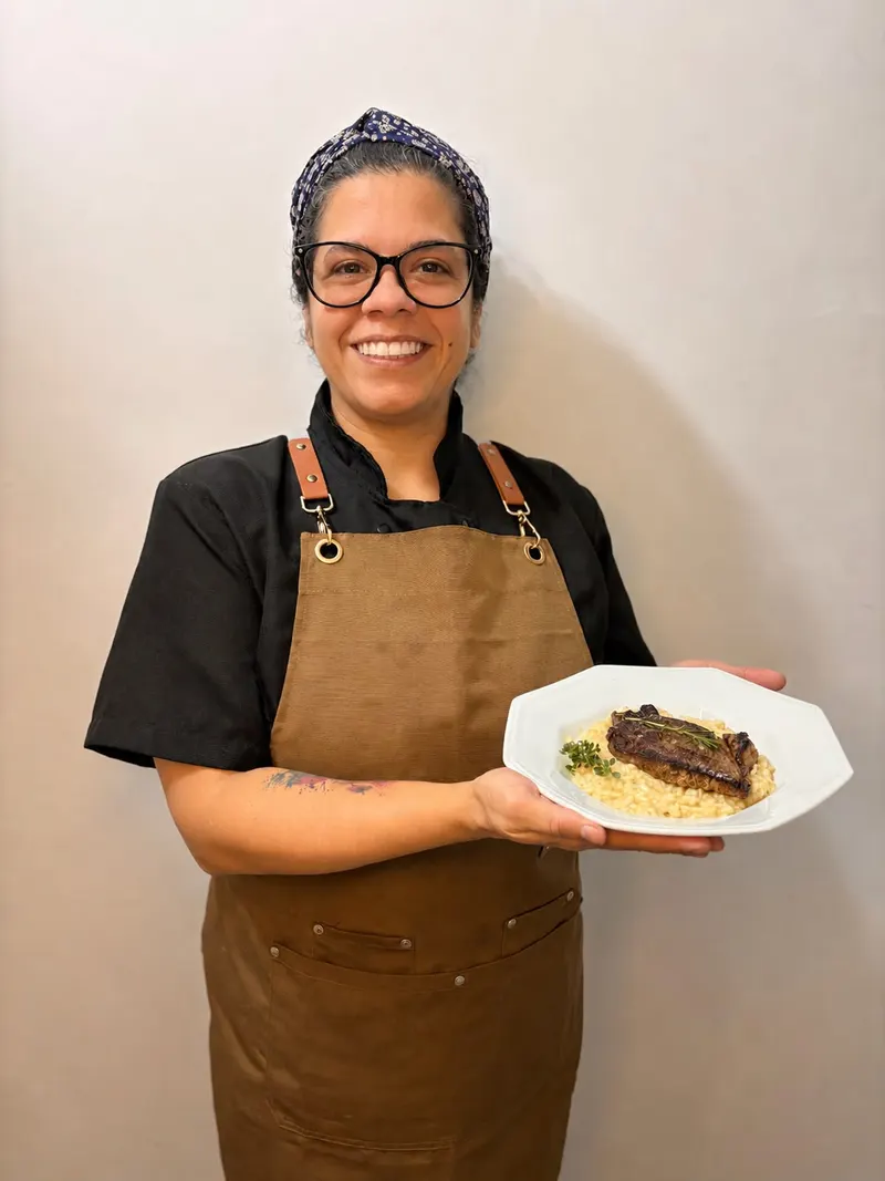 Chef Waléria Marques sorrindo, de dólmã e avental, segurando um prato de bife com risoto